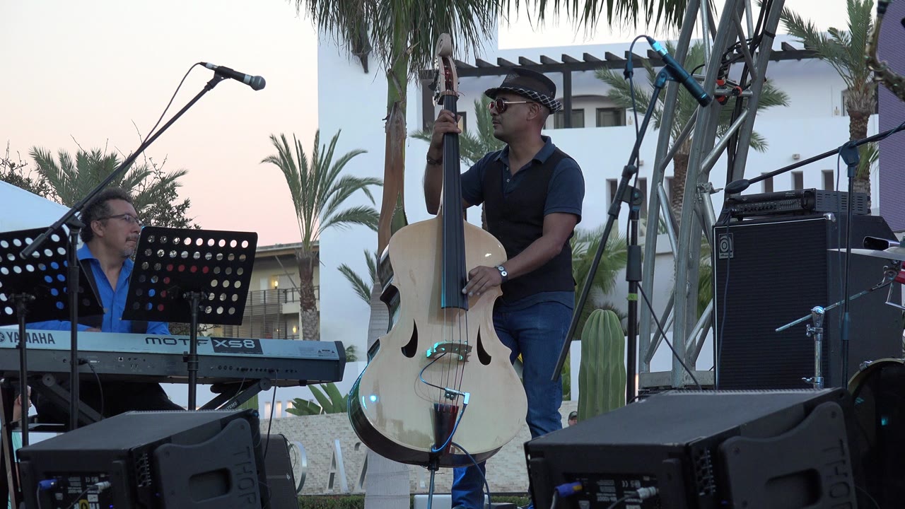 In Los Cabos, Mexico, the bass player stands more prominently with one hand on the neck of the white upright bass as the keyboardist remains seated behind a perforated music stand.