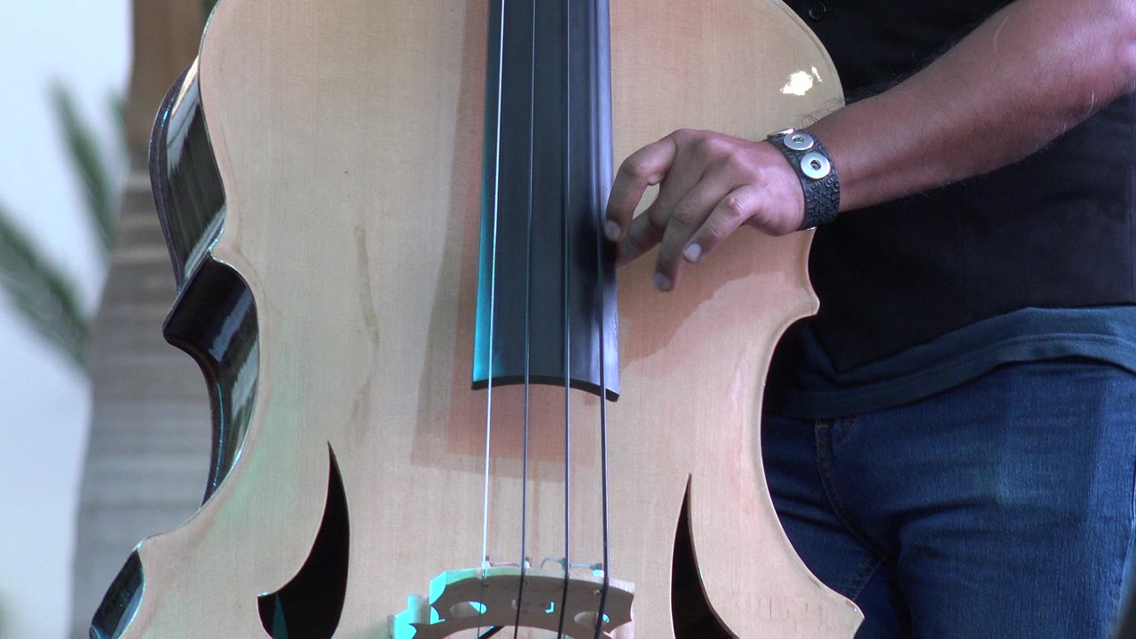 At 0:00 in Los Cabos, Mexico, a cropped adult man in a black T-shirt stands beside a pale wood cello, with his hand near the fingerboard and the body filling the frame.