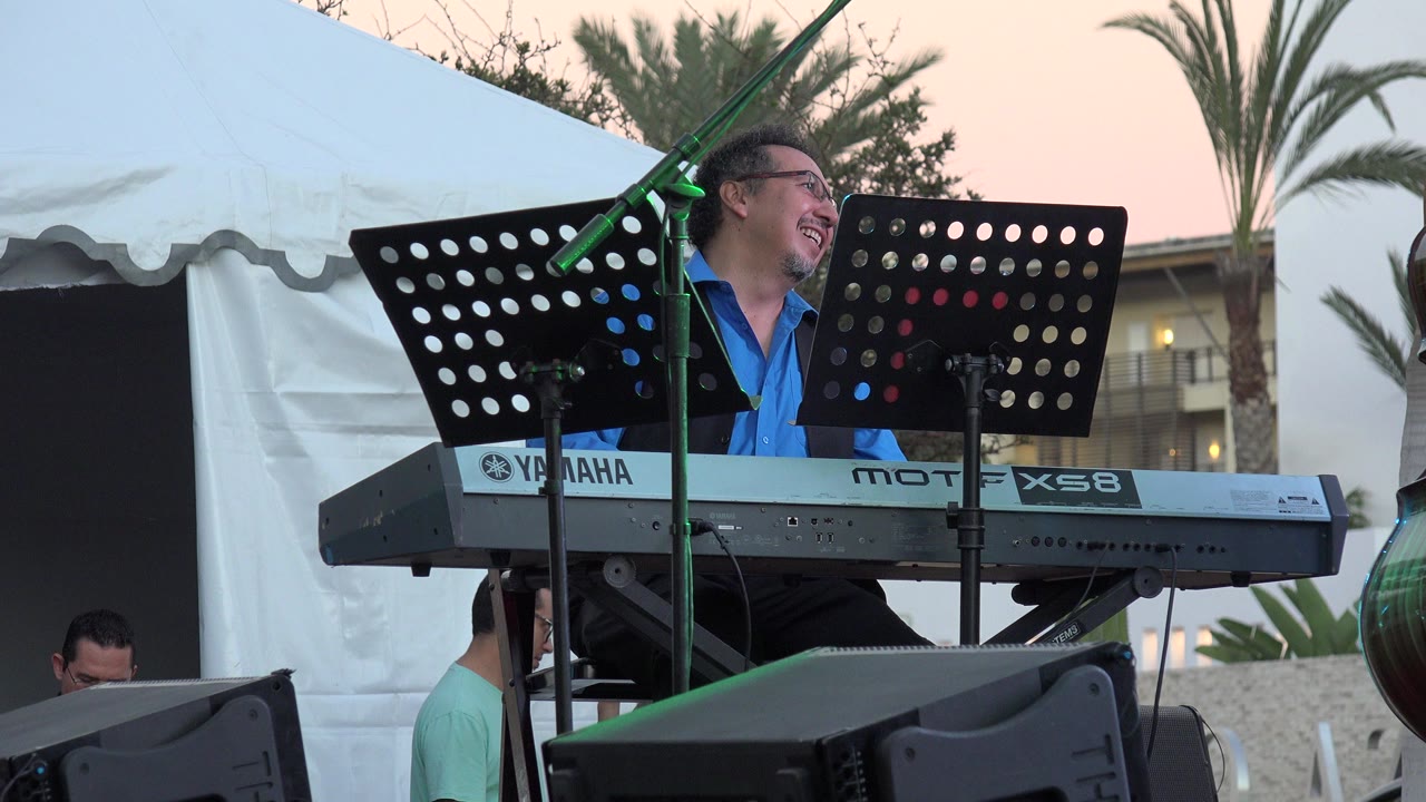 Keyboardist smiling behind a Yamaha keyboard and black music stands outdoors.