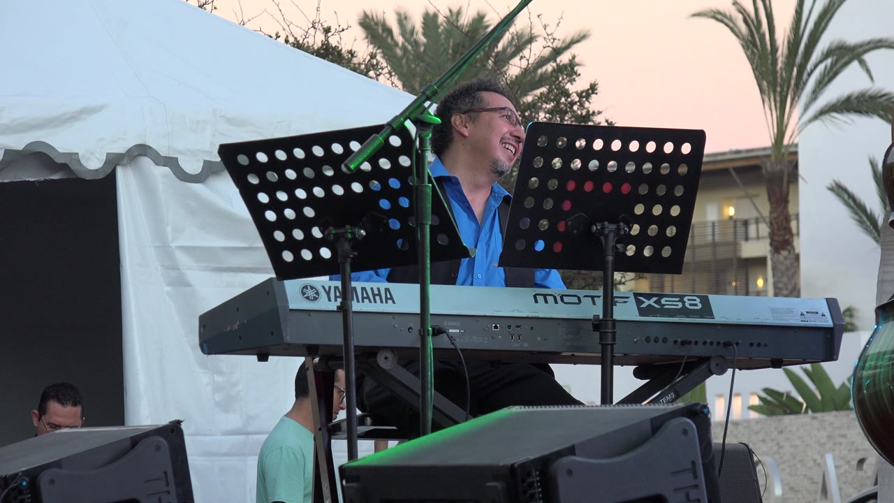 The keyboardist continues laughing behind the perforated music stands, with the Yamaha keyboard across the frame and Los Cabos palms and tent fabric visible behind the stage.