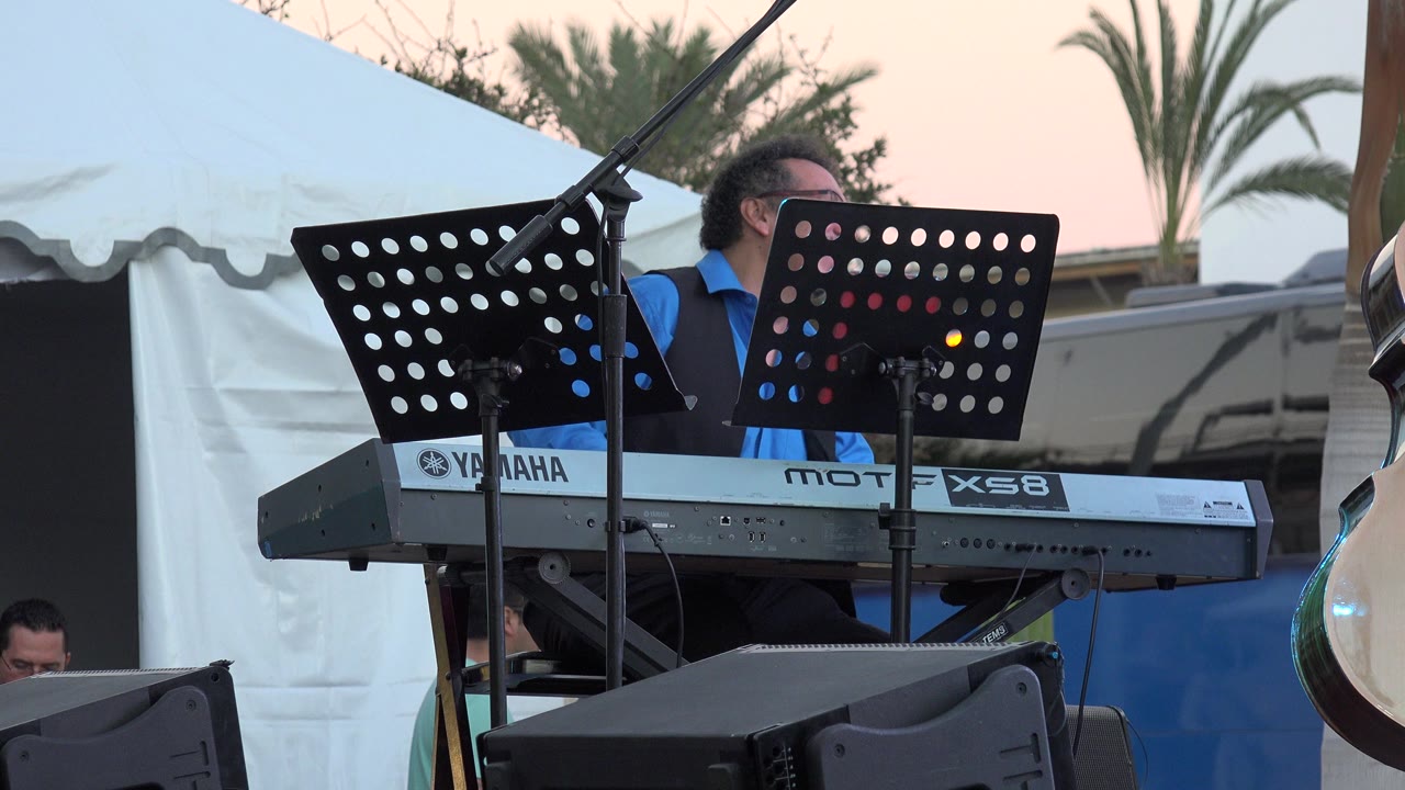 In Los Cabos, Mexico, a man in glasses sits behind a Yamaha keyboard while black music stands and a microphone boom fill the foreground.