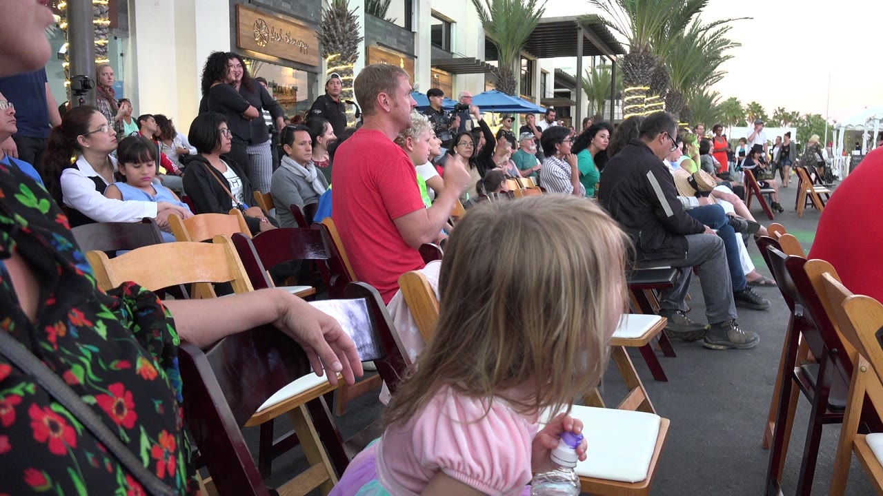 Seated audience with children on wooden chairs outdoors.