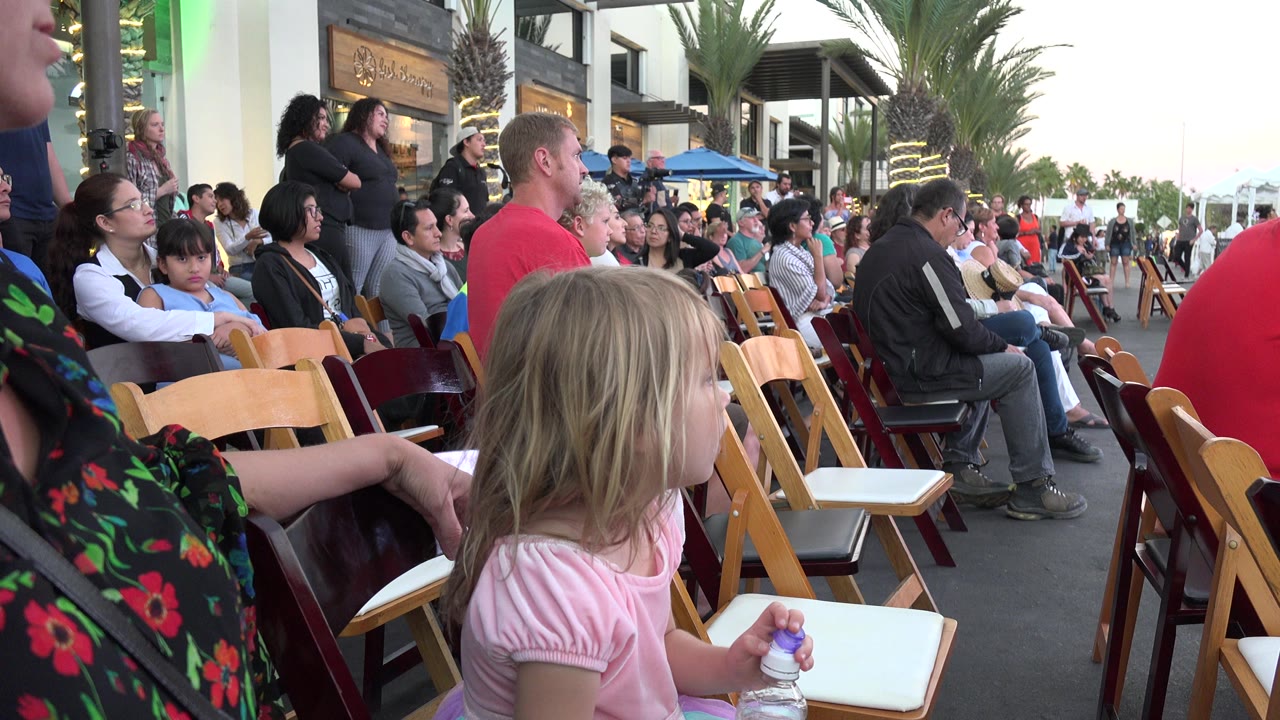 Young girl holding a water bottle beside rows of chairs at an outdoor audience area.