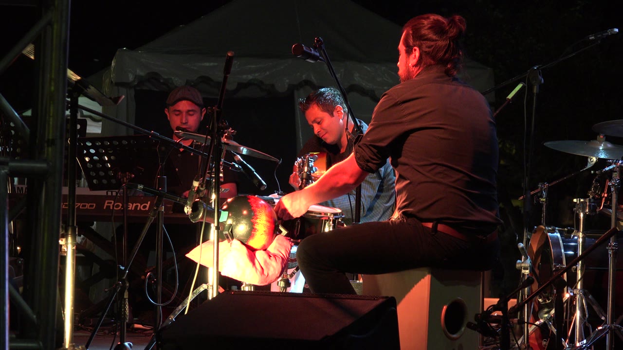 At 0:00 in Los Cabos, Mexico, three adult musicians sit among a keyboard, guitar, drums, microphones, and a music stand under warm red stage lighting.