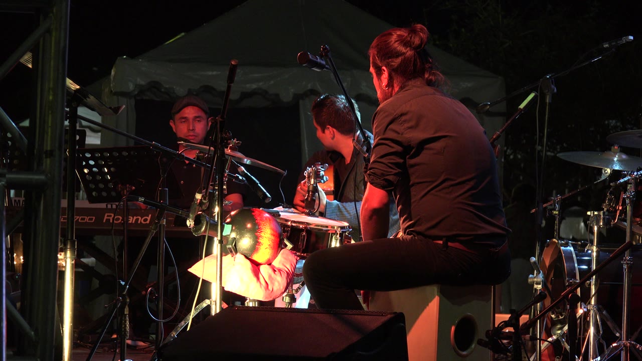 At 0:00 in Los Cabos, Mexico, musicians perform on a small stage with a foreground percussionist, guitar player, keyboard, microphones, and warm red lighting.