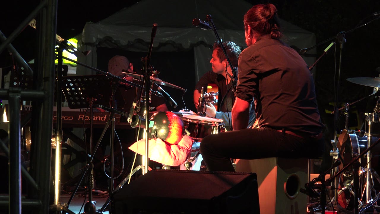 In Los Cabos, Mexico, a man in a black shirt sits on a tan speaker or box and taps a hand drum while another musician and stage equipment sit behind him.