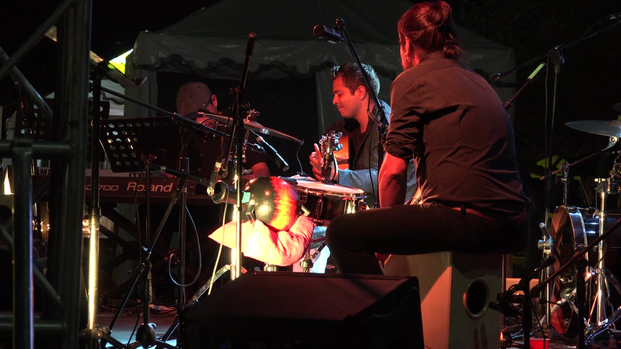 Musicians sit among drums, cymbals, cables, and microphones.