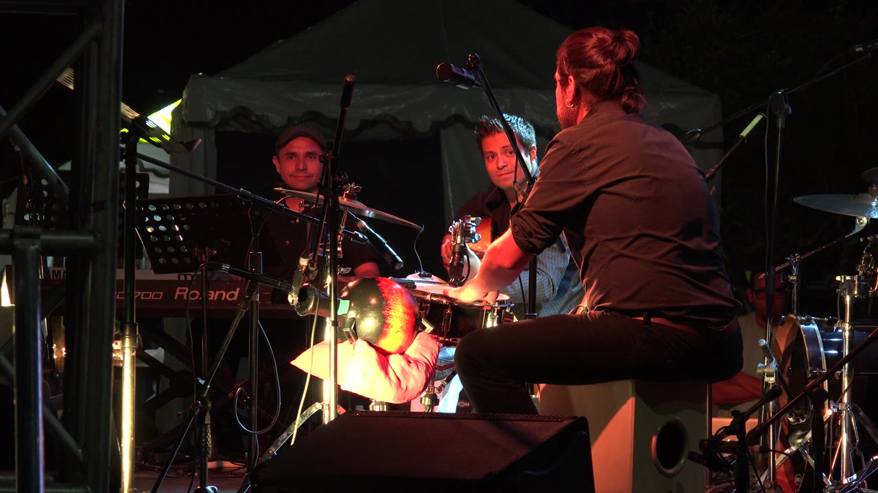 In Los Cabos, Mexico, a seated percussionist faces two musicians on a dark outdoor stage with microphones, keyboard, drums, and red stage light.