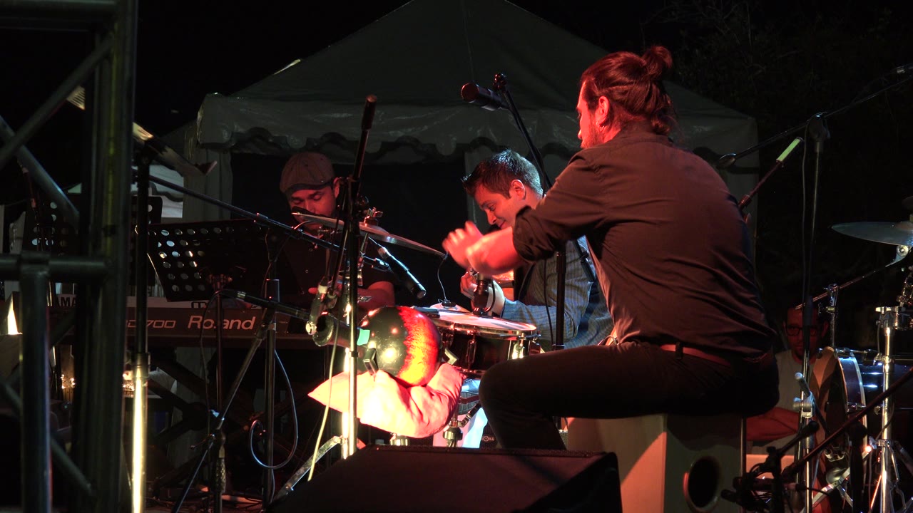 At 0:01, the foreground percussionist leans over the hand drum while the guitarist looks down at his instrument and the keyboard player remains partly visible at left in Los Cabos, Mexico.