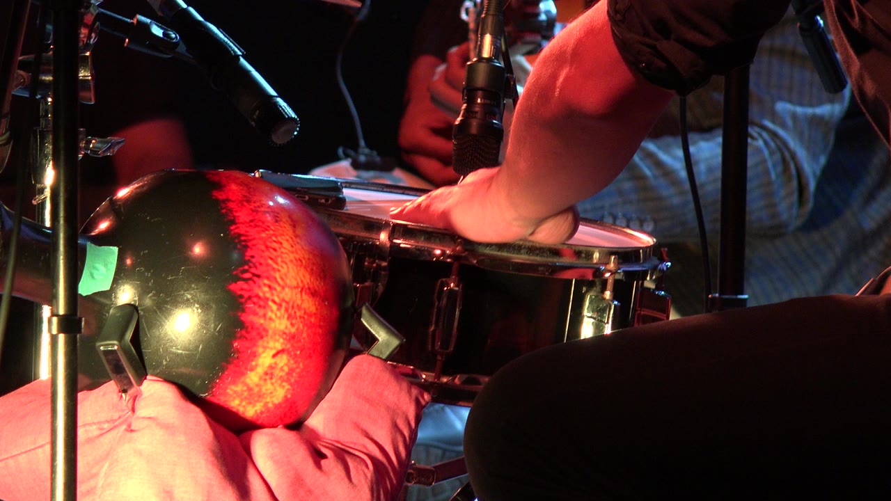 Close-up in Los Cabos, Mexico of a hand on a small drum beside a glossy red and black percussion instrument, with microphone stands and partial musicians visible.
