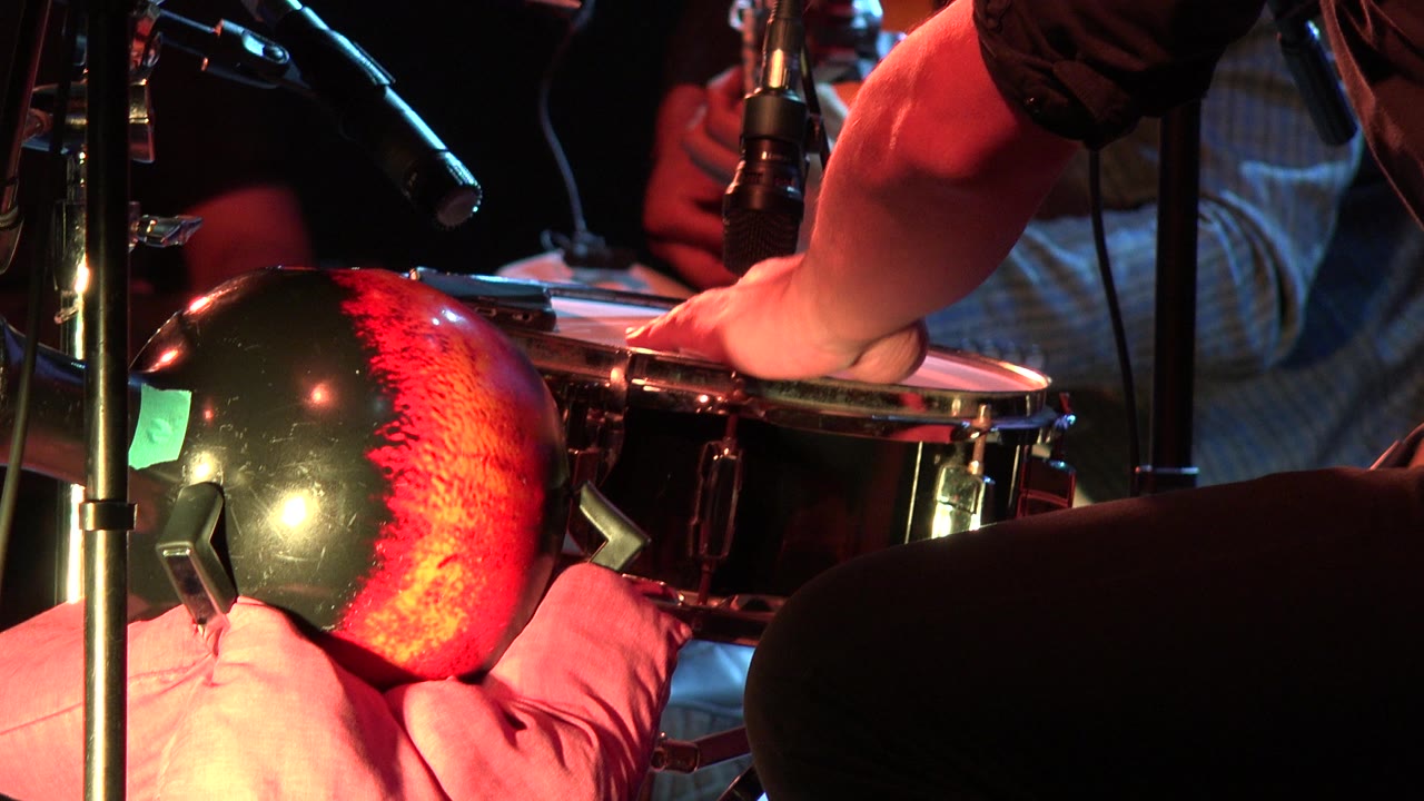 A musician's hand rests on the drumhead in Los Cabos, Mexico, framed by a red and black percussion instrument, mic stands, cables, and a partially seen guitar player.