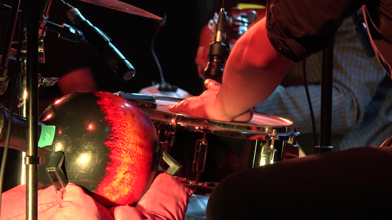 A close Los Cabos performance frame shows the drummer's forearm stretched across the snare, with overhead microphones and warm red light on the drum kit.