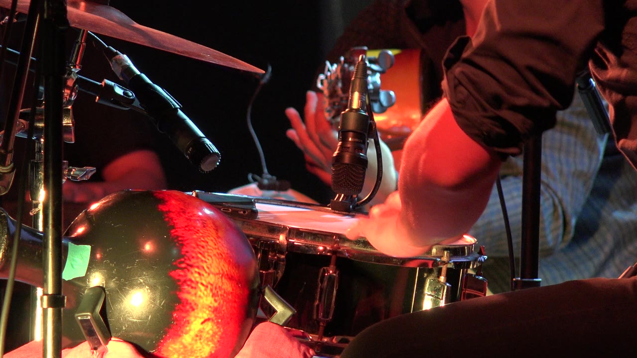 At 0:01, the Los Cabos Mexico footage holds close on the snare as a hand lands near the drumhead rim, with microphones and cymbal hardware surrounding it.