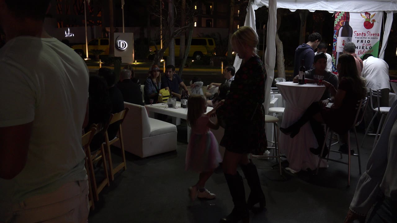 At 0:01 in Los Cabos, the girl in the pink dress faces the woman beside a high table, with diners seated under a white canopy and street signage visible behind them.