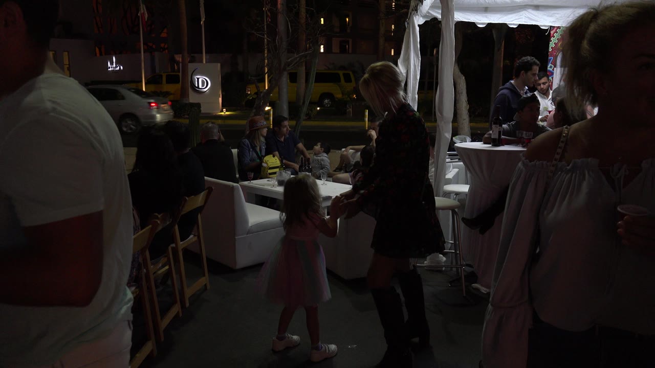 In Los Cabos, Mexico, adults stand near white lounge furniture while a young girl in a pink dress is visible in the darker midground at an outdoor nighttime gathering.