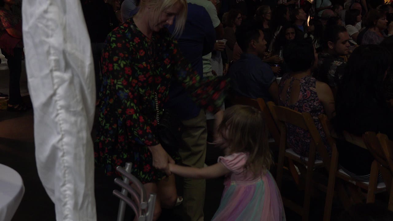 Adult woman holding a young girl's hand beside seated event guests.
