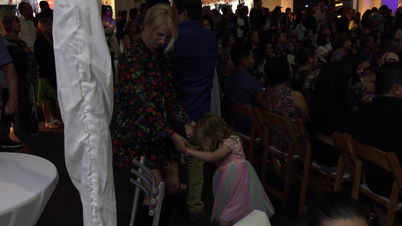 The woman and child remain hand in hand at the Los Cabos gathering, framed by a white pole in front and rows of seated people behind them.