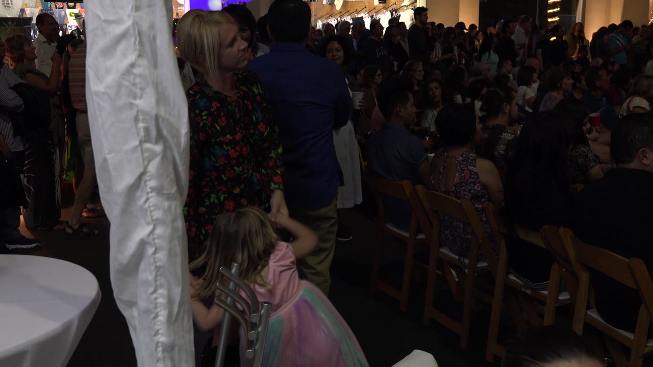 The woman remains beside the little girl near the white column while a dense crowd of seated and standing guests fills the Los Cabos party background.