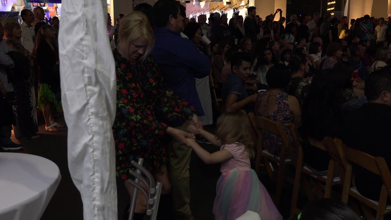 At a Los Cabos wedding reception, a woman in a dark floral dress holds a young child's hand beside chairs, tables, and seated guests in dim event light.