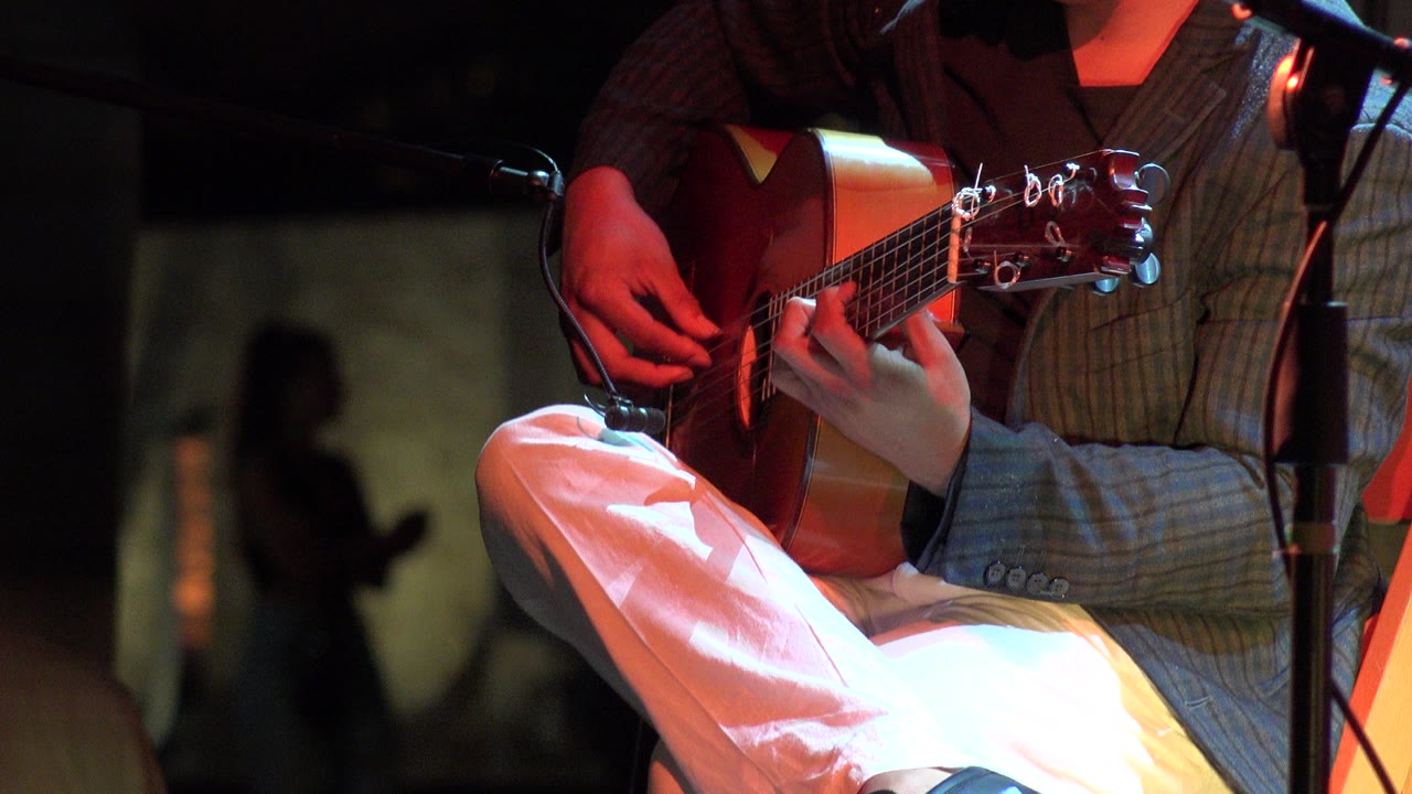 Close framing shows a man in white pants and a plaid jacket playing classical guitar on a small stage in Los Cabos, Mexico, with a microphone stand at the left edge.