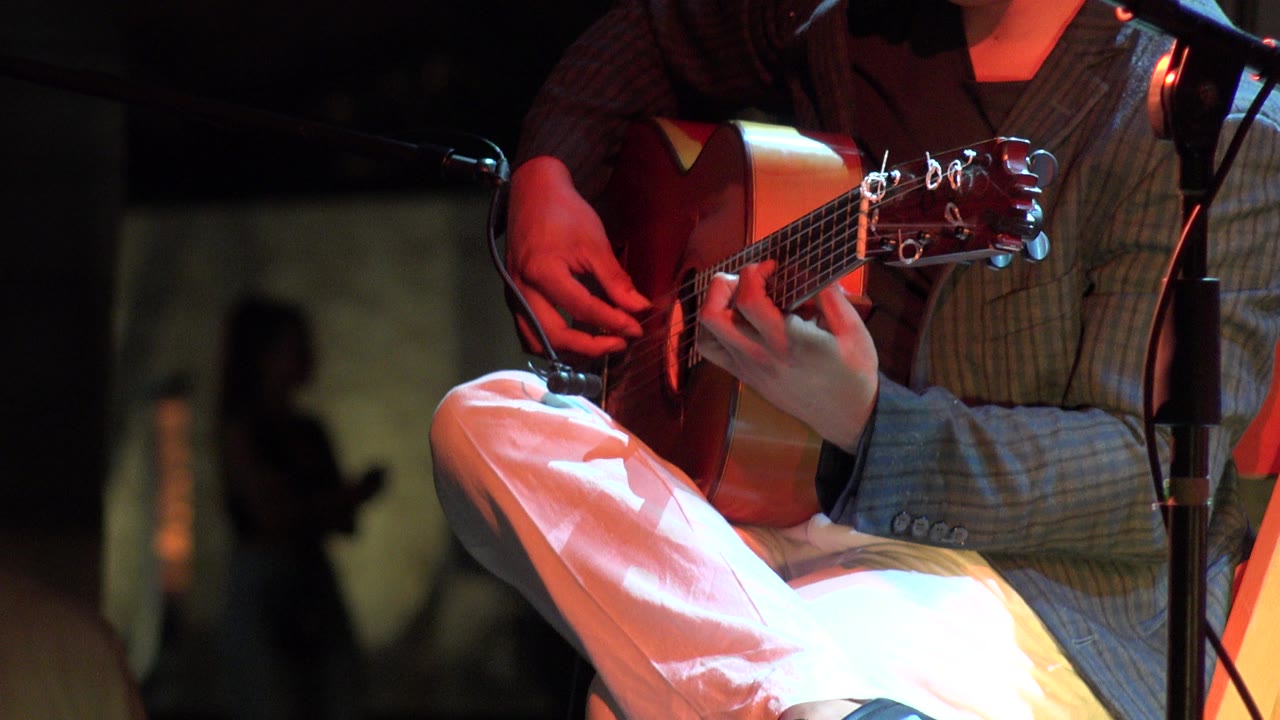 The musician's picking hand and fretting hand are centered on the classical guitar, with red stage lighting and a blurred standing figure in the background.