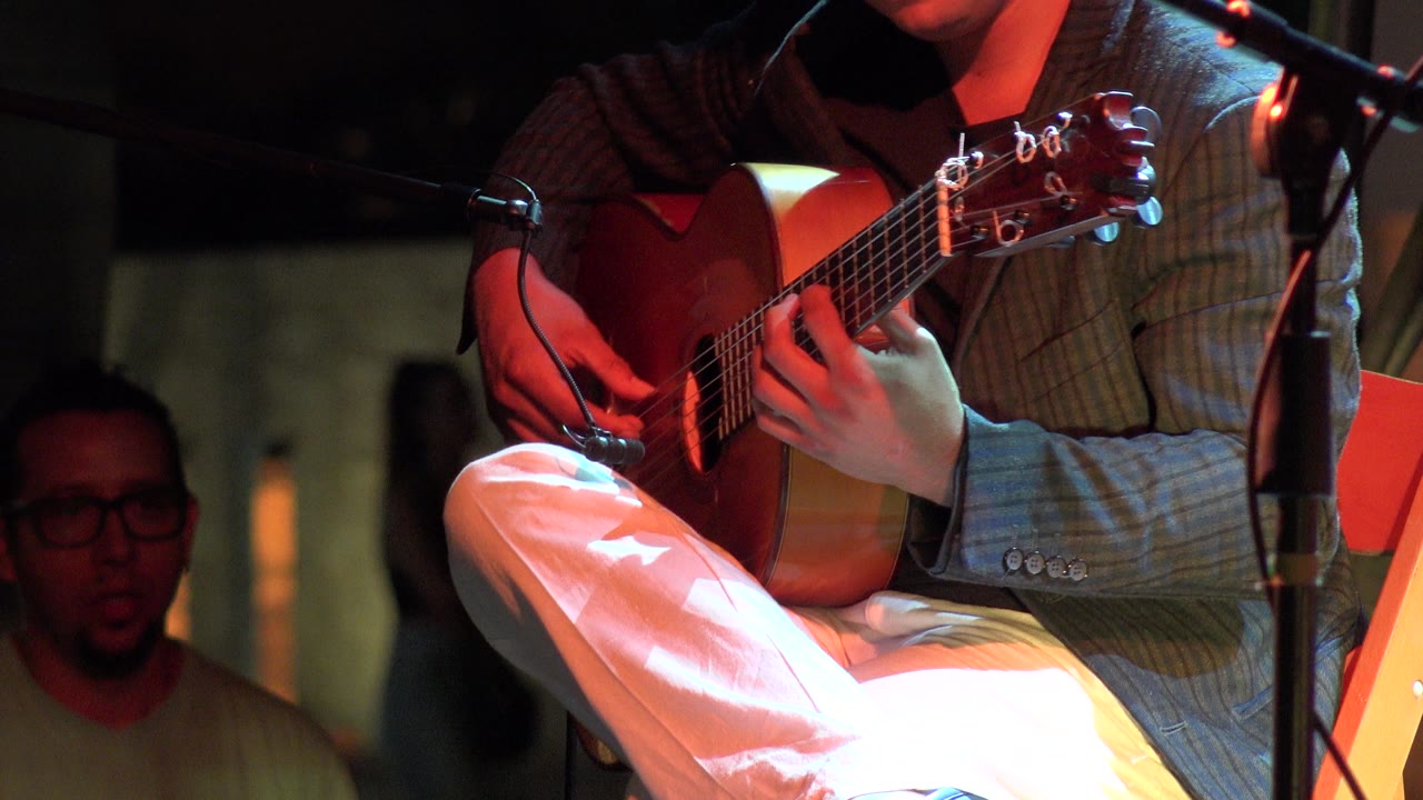 Seated guitarist with microphone stands and background attendee.