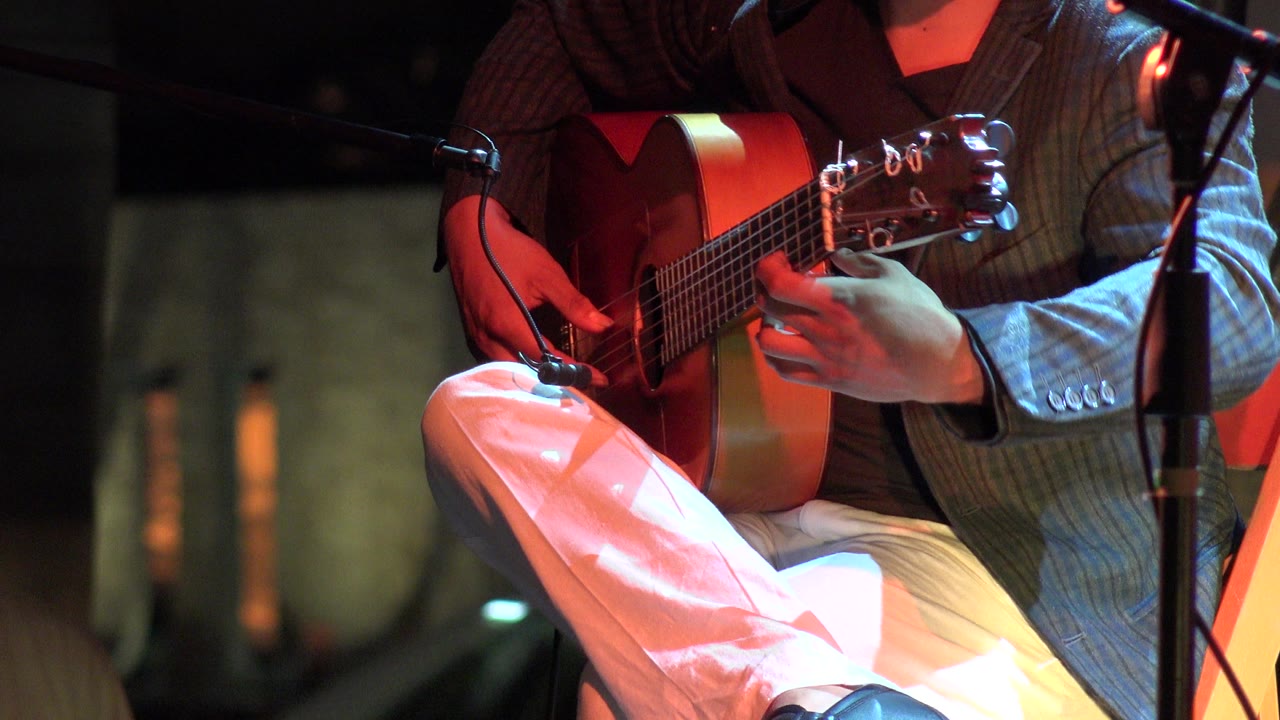 Close frame of a seated musician in Los Cabos, Mexico playing an acoustic guitar, with the guitar body, picking hand, white pants, and a microphone stand visible.