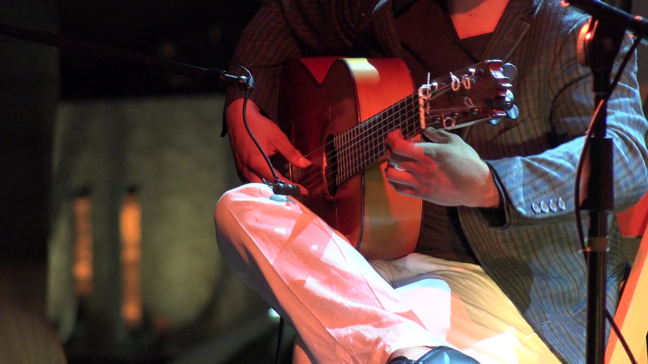 A close Los Cabos restaurant performance frame shows the musician's fretting hand near the guitar headstock and a black microphone stand crossing the foreground.