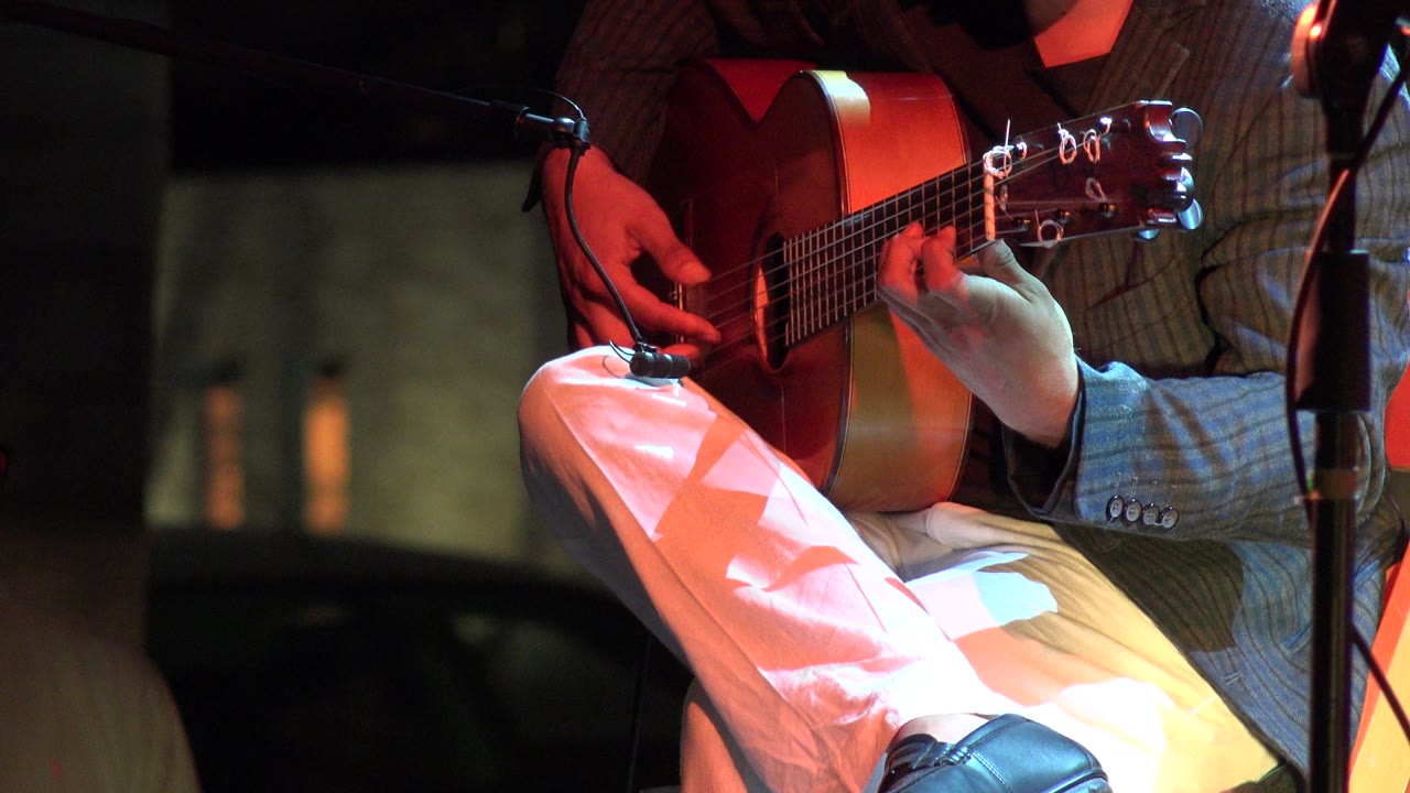 The Los Cabos house show close-up holds on the acoustic guitar as the performer's picking hand rests near the sound hole and the microphone stand sits at frame right.