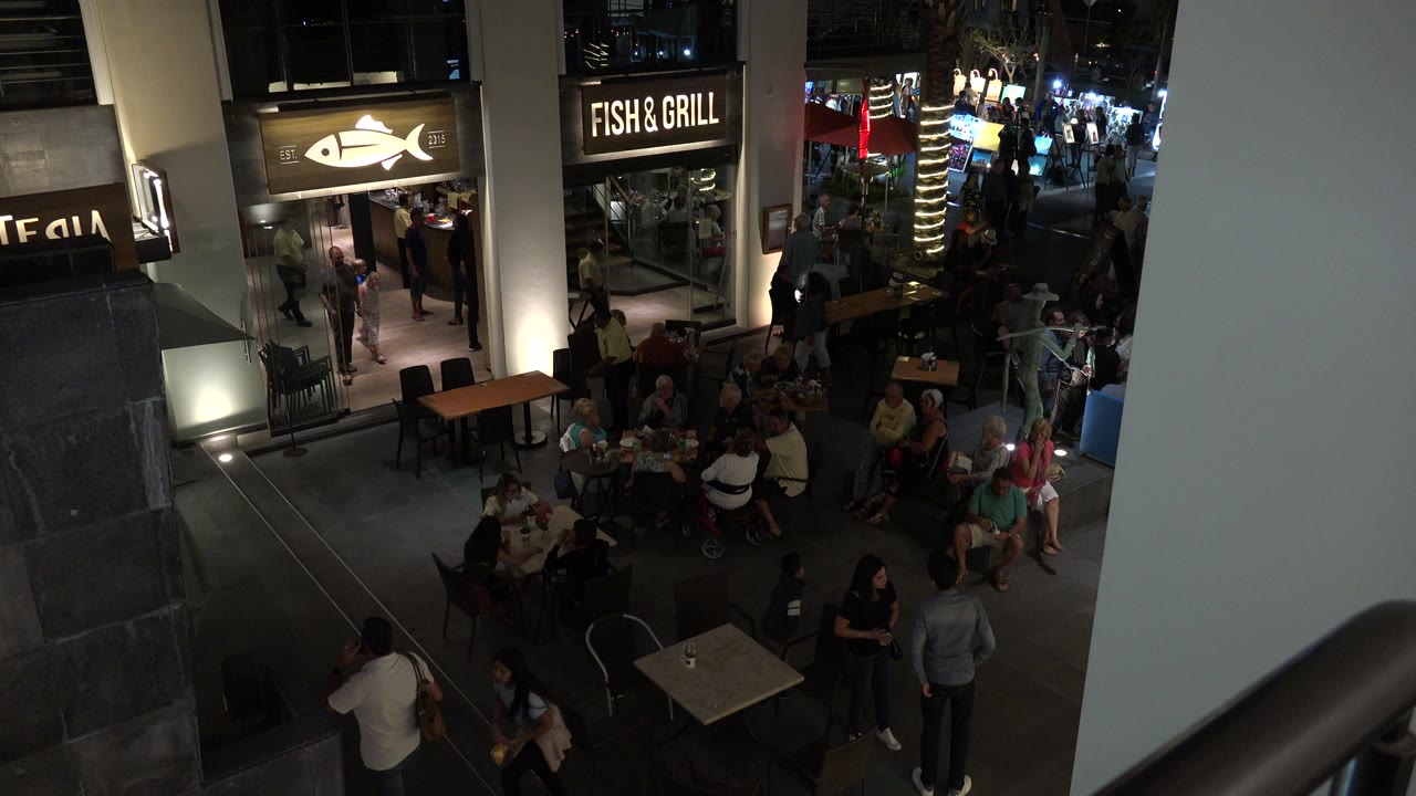 Los Cabos mall corridor with seated diners, pedestrians, lit restaurant signs, and a pale foreground wall framing the right side one second in.