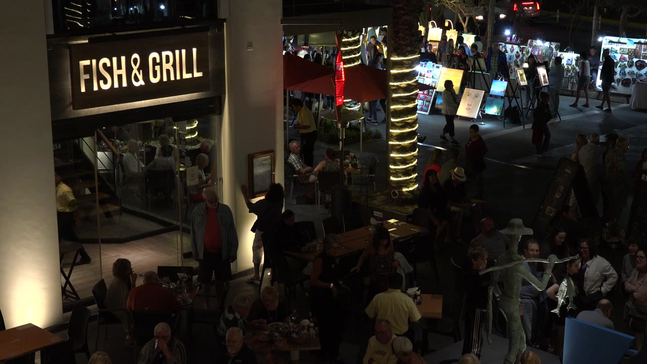At 0:01, the Los Cabos food court view shows diners seated in the lower frame, a glowing restaurant sign on the left, and people moving along the walkway in the background.