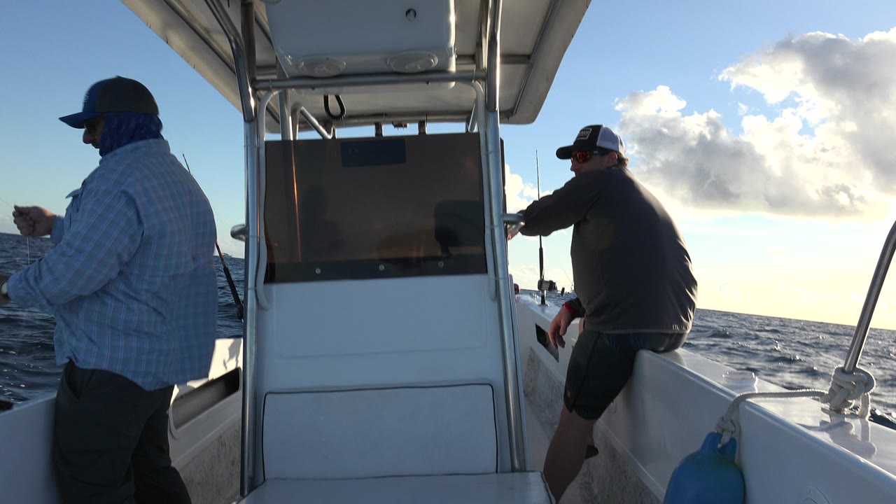 The left man stands near the rail while the right man leans over the side of the boat, with dark water and bright cloud edges behind them in Los Cabos, Mexico.