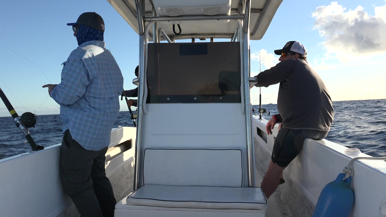 The standing angler in a light shirt remains on the left side of the boat while another man leans on the right rail, with Los Cabos open water behind them.