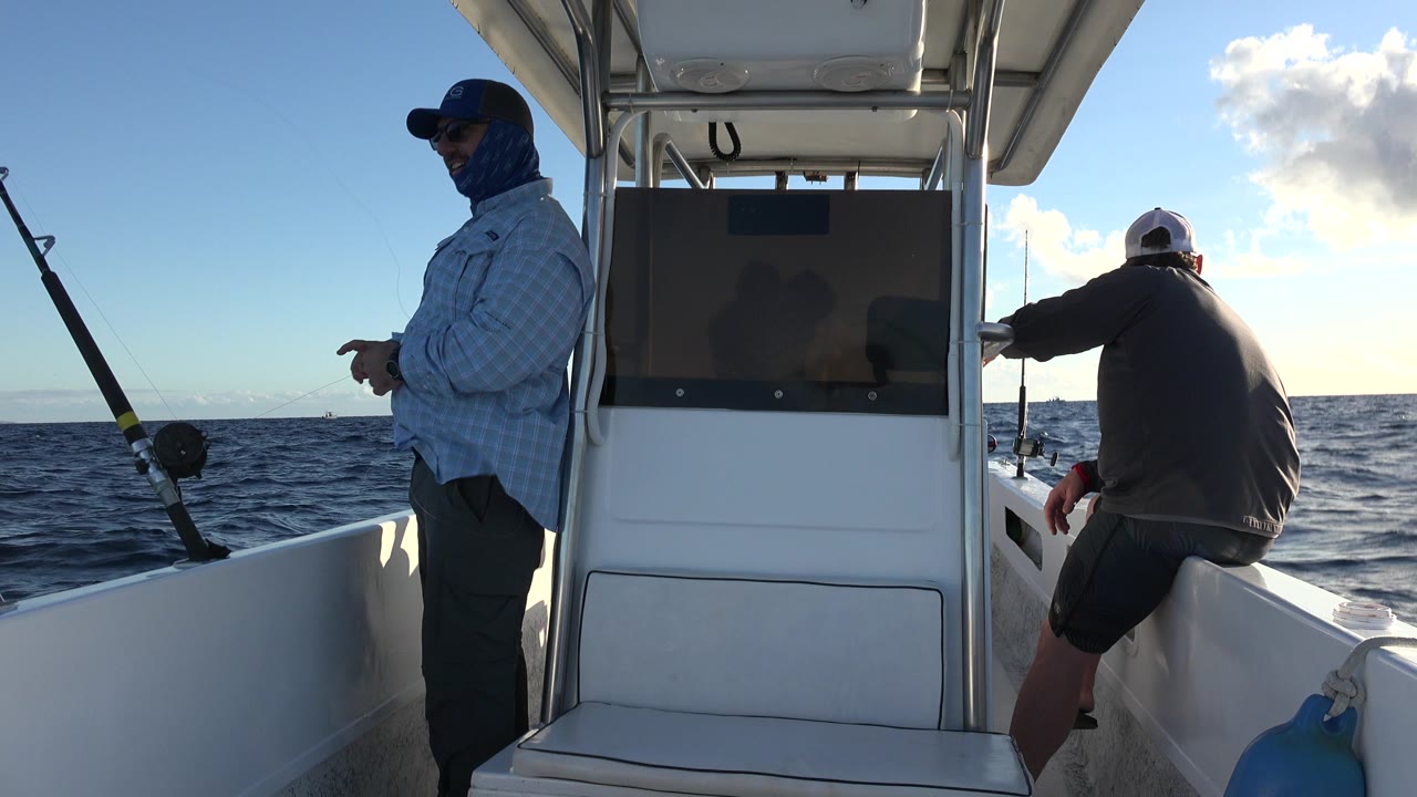 Two adult men are on a small white fishing boat off Los Cabos, Mexico, with one standing by a rod at left, another seated at right, and low sunlight over the ocean horizon.