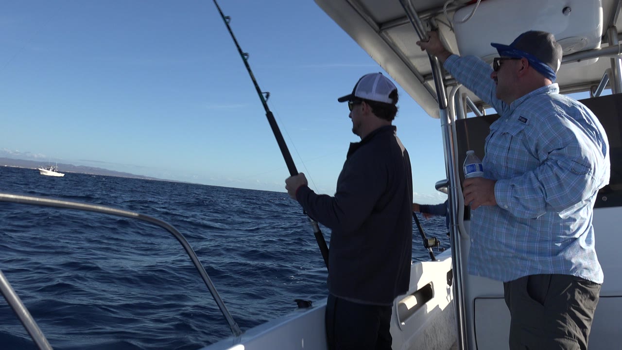 At 0:00 in Los Cabos, Mexico, two adult men stand on a small boat, with one holding a fishing rod beside the rail and distant boats on the horizon.