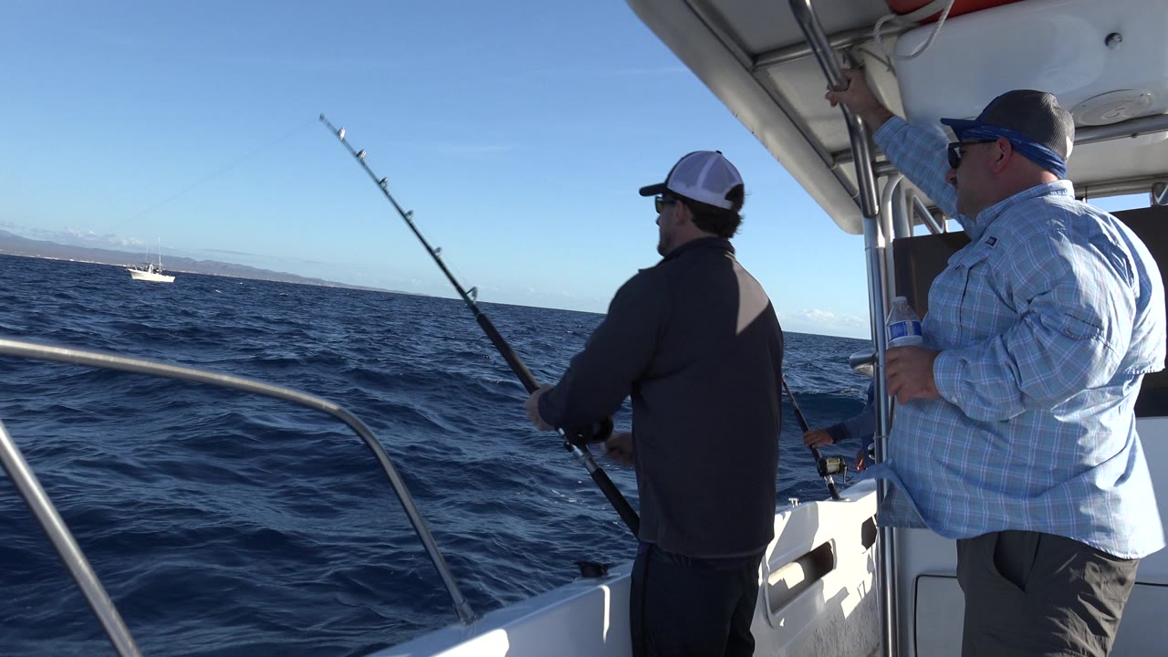 Man reels a fishing rod from a small boat as another man stands under the canopy.