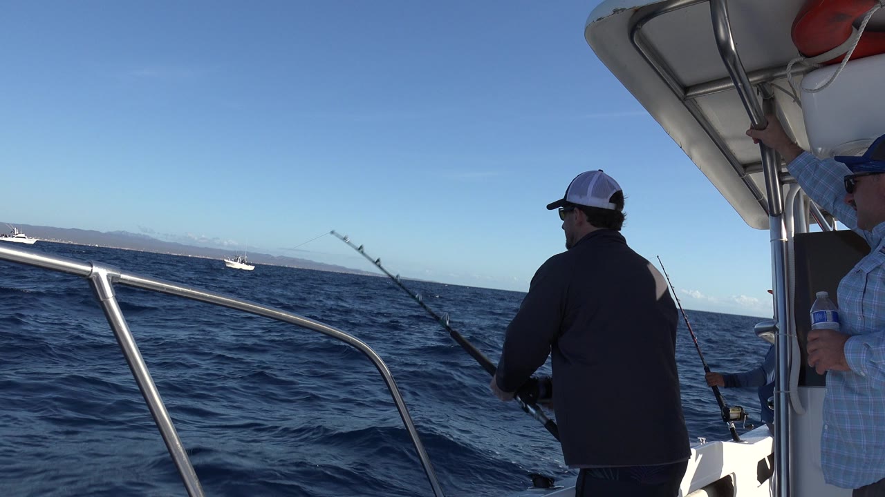 A man in a cap fishes from a boat off Los Cabos, with metal railings in the foreground, dark blue waves, distant land, and small boats on the horizon.