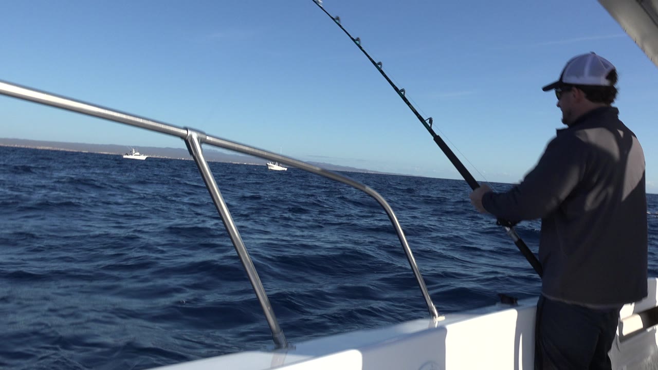 The fishing rod angles upward beside the boat rail as the man faces the water, with two small boats visible beyond the rolling sea in Los Cabos Mexico.
