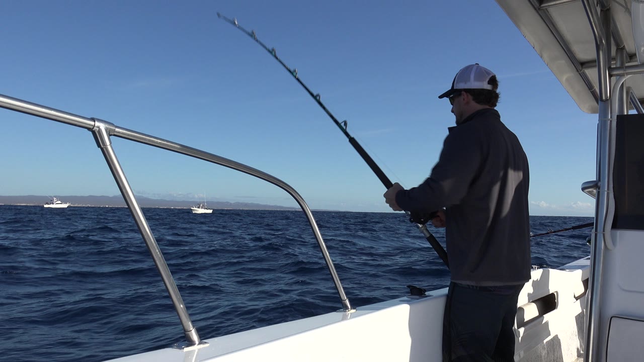 At the start of the Los Cabos fishing clip, a man stands at the right side of a white boat with a raised rod over dark blue water and distant boats on the horizon.
