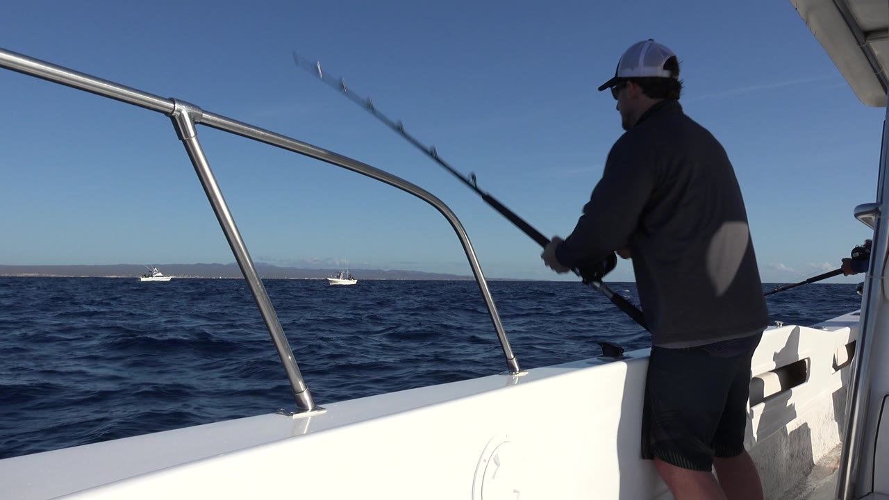 At 0:00, a man in a gray jacket and white cap fishes from a white boat in Los Cabos, Mexico, with the boat rail crossing the foreground and small boats on the horizon.