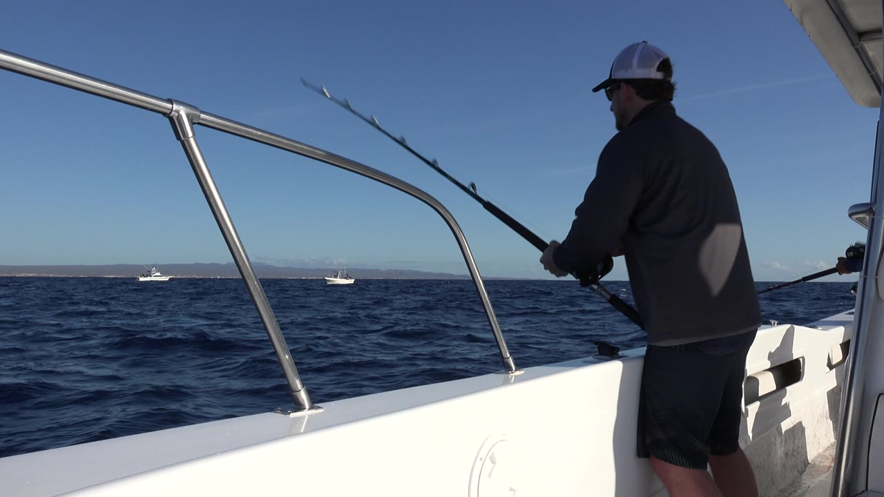 At 0:01, the man remains near the right side of the frame holding the black fishing rod, with the white boat rail, blue water, distant boats, and pale coastline visible.