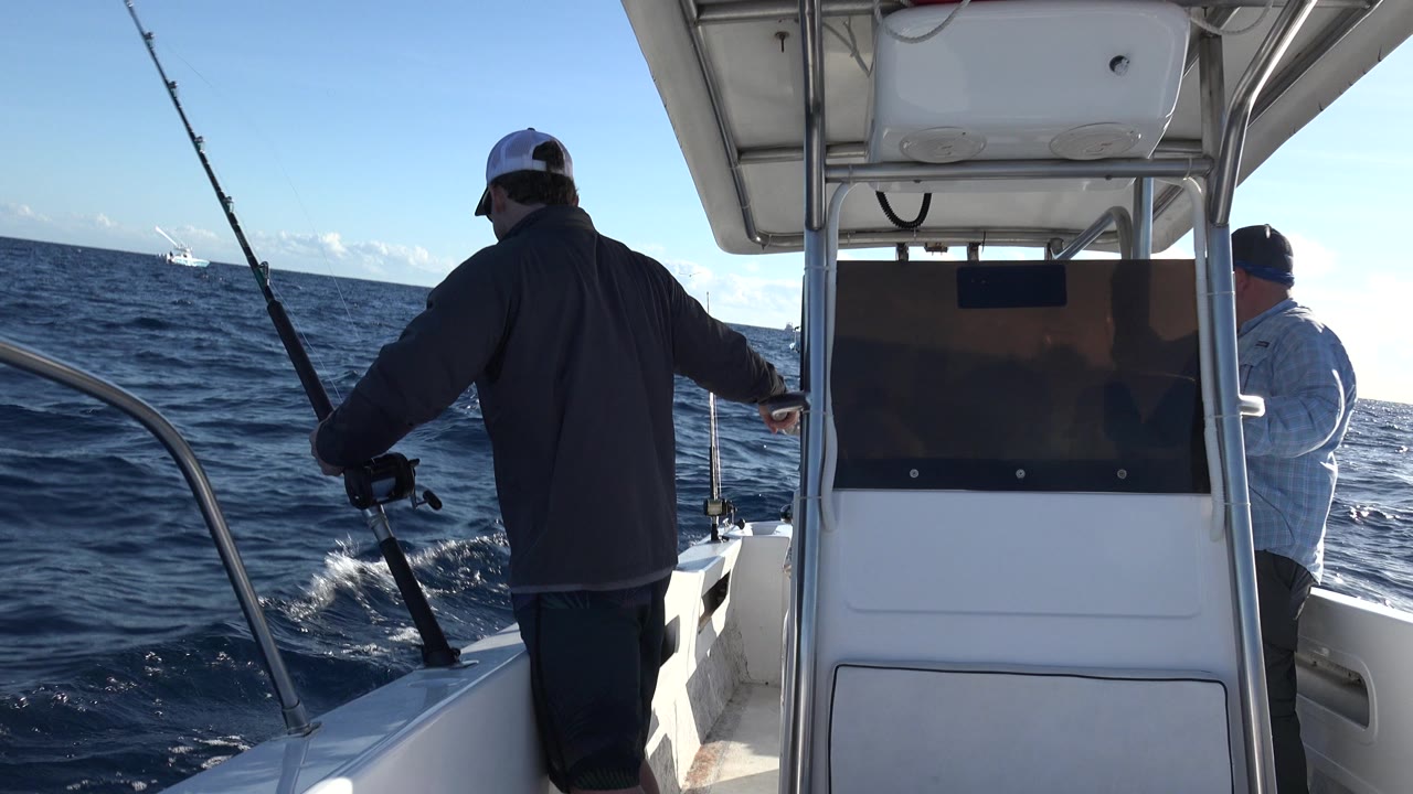 Two men stand on a white boat in Los Cabos, Mexico, one holding a black fishing pole near the rail and the other facing the water beside the center console.