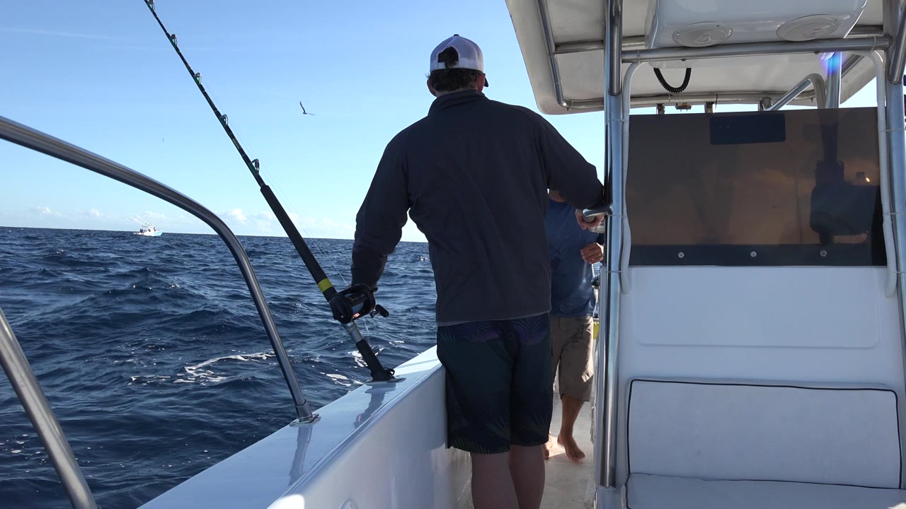 A man in a cap faces away on the boat deck in Los Cabos, Mexico, while another person is partly visible near the console and fishing rods frame the ocean view.