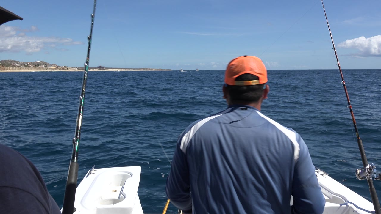 The man remains centered from behind on the Los Cabos fishing boat, with two rods rising through the frame and a low shoreline across the water.