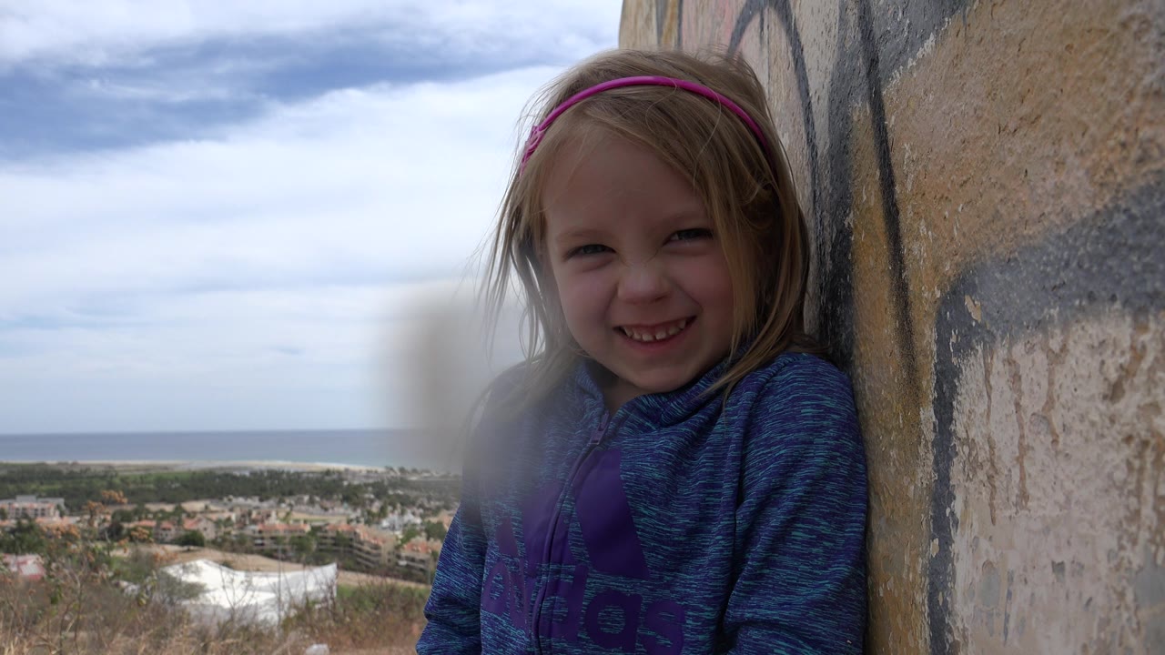One second in, the girl remains close to the painted wall, smiling toward the camera with the Los Cabos townscape and ocean horizon visible over the hillside.