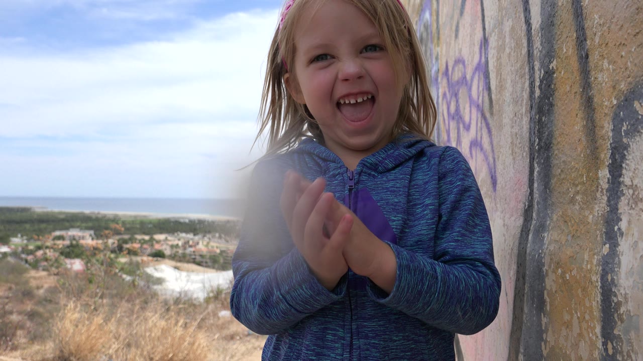 The girl faces the camera with her hands together beside the marked wall, while the Los Cabos hillside, town area, ocean line, and pale cloudy sky remain visible behind her.