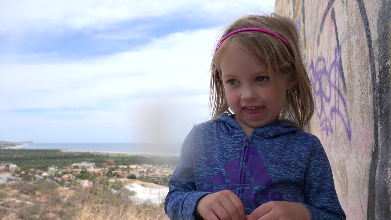 Young child in a blue hoodie beside a marked wall with ocean and town below.