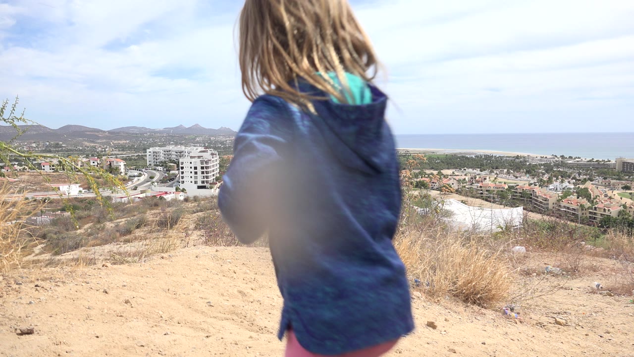 Young girl in blue hoodie on dry hillside above coastal buildings and ocean.