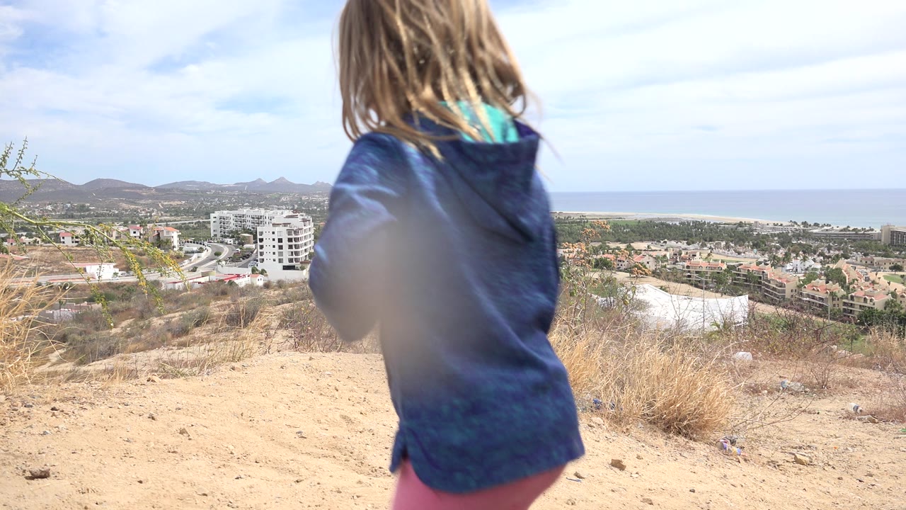 The girl remains close to the low camera on a dusty Los Cabos hillside, with scrub brush, white buildings, distant hills, and the ocean horizon visible under bright daylight.