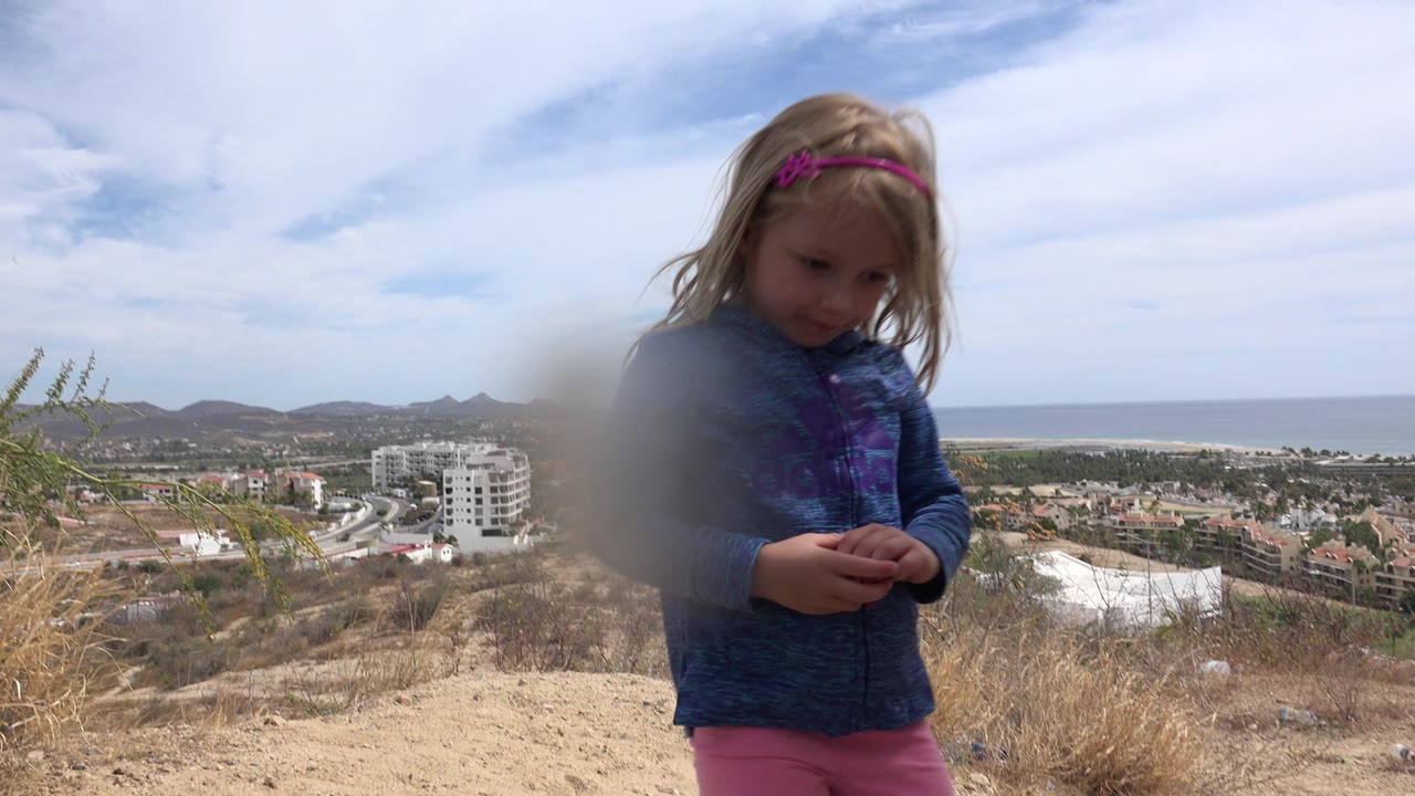 In Los Cabos, Mexico, the girl remains near the foreground, looking down at her hands as the ocean horizon and hillside buildings sit behind her.